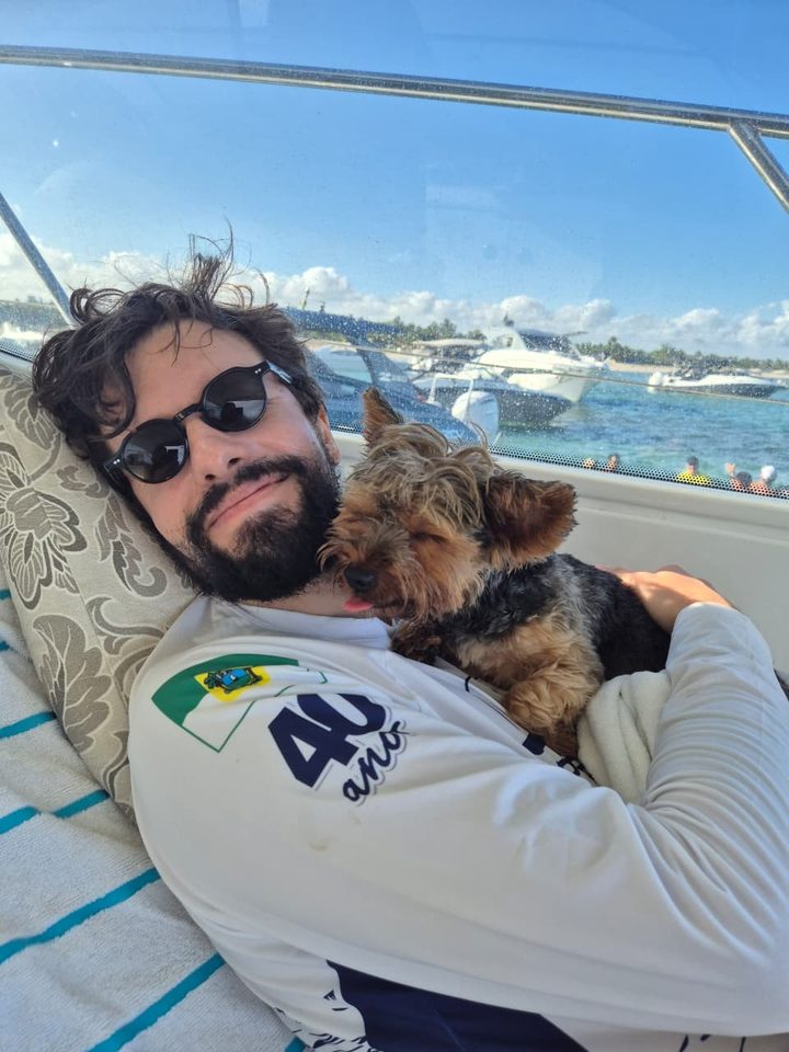 Luccas de Amorim num barco, de óculos escuros, abraçado à sua cachorra, uma yorkshire; ao fundo, o mar e outros barcos.